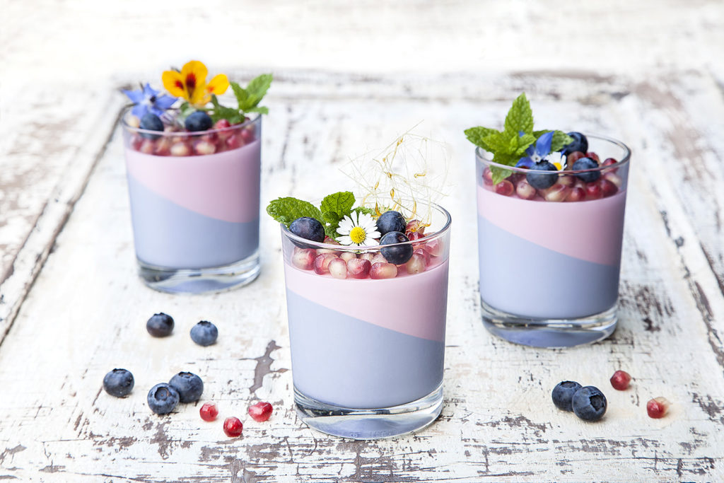 Professionelle Foodfotografie in Bielefeld – Dessert im Glas mit flieder- und rosafarbenen Schichten, dekoriert mit Beeren, Karamell und Blumen